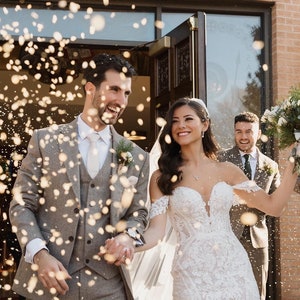 Peut inclure: Une mari&eacute;e et un mari&eacute; sortent d'une &eacute;glise, souriants et se tenant la main, tandis que des invit&eacute;s lancent des confettis. La mari&eacute;e porte une robe de mari&eacute;e blanche &agrave; &eacute;paules d&eacute;nud&eacute;es avec des d&eacute;tails en dentelle. Le mari&eacute; porte un costume gris avec une chemise blanche et une cravate.