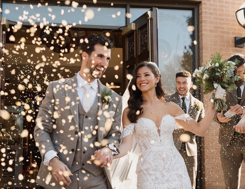 Peut inclure: Une mari&eacute;e et un mari&eacute; sortent d'une &eacute;glise, souriants et se tenant la main, tandis que des invit&eacute;s lancent des confettis. La mari&eacute;e porte une robe de mari&eacute;e blanche &agrave; &eacute;paules d&eacute;nud&eacute;es avec des d&eacute;tails en dentelle. Le mari&eacute; porte un costume gris avec une chemise blanche et une cravate.