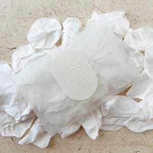May include: A collection of white tissue paper petals and a package wrapped in translucent paper. A white oval tag with the words "TOSS WITH LOVE" is on top of the package. The background is a light beige marble surface.