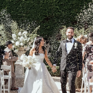 Peut inclure: Une mari&eacute;e et un mari&eacute; marchent dans l'all&eacute;e apr&egrave;s leur c&eacute;r&eacute;monie de mariage, arros&eacute;s de confettis. La mari&eacute;e porte une robe blanche sans bretelles et le mari&eacute; porte un smoking noir. Ils sourient tous les deux et se tiennent la main.