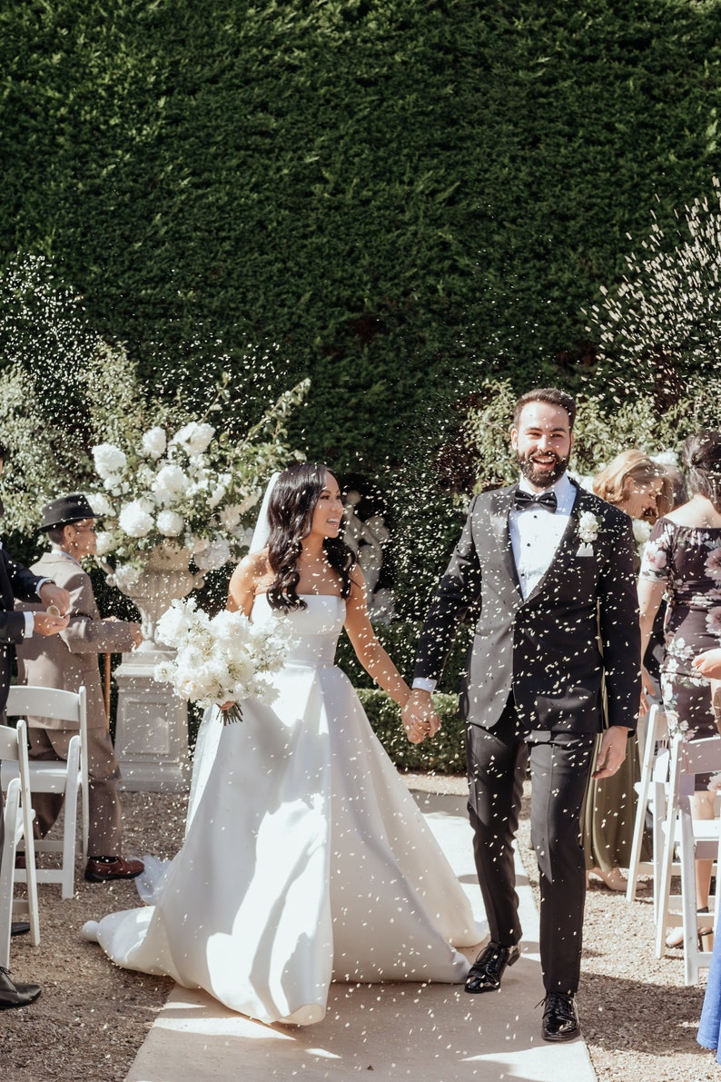 Peut inclure: Une mari&eacute;e et un mari&eacute; marchent dans l'all&eacute;e apr&egrave;s leur c&eacute;r&eacute;monie de mariage, arros&eacute;s de confettis. La mari&eacute;e porte une robe blanche sans bretelles et le mari&eacute; porte un smoking noir. Ils sourient tous les deux et se tiennent la main.