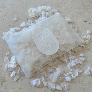Könnte beinhalten: Ein durchsichtiges Päckchen gefüllt mit weißen Blütenblättern, mit einem weißen ovalen Aufkleber mit der Aufschrift "TOSS WITH LOVE". Das Päckchen ist von verstreuten Blütenblättern umgeben, was eine zarte, romantische Ästhetik erzeugt.