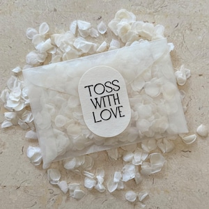 May include: A transparent cellophane packet filled with white flower petals, surrounded by scattered petals. The packet has a white oval sticker with the text "TOSS WITH LOVE" in black. The background is a light beige surface.
