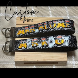 May include: Two black wrist keychains with the names "MRS BLUME" and "MRS ROOT" in graffiti-style lettering. The text is outlined in white with orange and yellow shading. Each keychain has a silver-colored metal clasp.