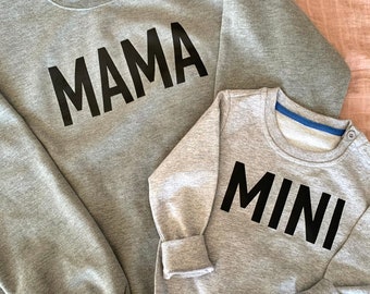 May include: Two gray sweatshirts with black text. The larger sweatshirt says "MAMA" and the smaller sweatshirt says "MINI".