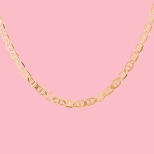 May include: A gold chain necklace with a mariner link design. The chain is hanging against a pink background.