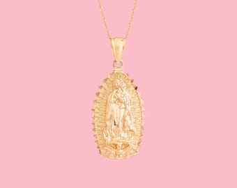 May include: A gold-colored pendant necklace featuring a detailed depiction of the Virgin Mary. The pendant is oval-shaped with a textured border and hangs from a delicate gold chain. The background is a solid pink.