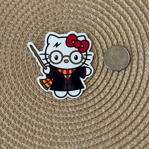 K&ouml;nnte beinhalten: Ein wei&szlig;er Hello Kitty-Aufkleber mit einer roten Schleife, der schwarze Roben, eine Brille und einen gelben und roten Schal tr&auml;gt. Sie h&auml;lt einen Zauberstab und hat einen Blitz auf der Stirn.