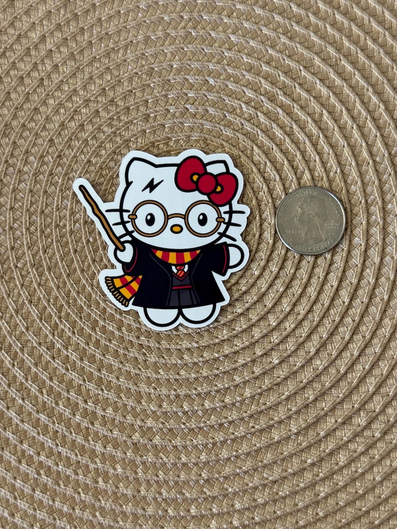 K&ouml;nnte beinhalten: Ein wei&szlig;er Hello Kitty-Aufkleber mit einer roten Schleife, der schwarze Roben, eine Brille und einen gelben und roten Schal tr&auml;gt. Sie h&auml;lt einen Zauberstab und hat einen Blitz auf der Stirn.