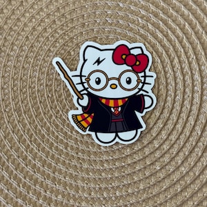 K&ouml;nnte beinhalten: Weisse Hello Kitty Figur mit schwarzem Umhang, roter Schleife und runden Brillen. Die Figur h&auml;lt einen Zauberstab und hat einen Blitz auf der Stirn.