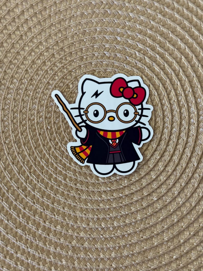 K&ouml;nnte beinhalten: Weisse Hello Kitty Figur mit schwarzem Umhang, roter Schleife und runden Brillen. Die Figur h&auml;lt einen Zauberstab und hat einen Blitz auf der Stirn.