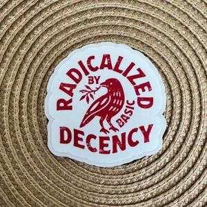 May include: A white sticker with a red design and text. The design features a bird holding a sprig, surrounded by the words "RADICALIZED BY BASIC DECENCY" in a circular arrangement. The sticker is on a woven, tan surface.
