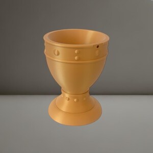 Könnte beinhalten: Ein goldfarbener, 3D-gedruckter Eierbecher. Der Becher hat einen abgerundeten Boden, einen Stiel mit dekorativen Nieten und eine breite, offene Oberseite. Das Design weist horizontale Bänder und Nietendetails auf und verleiht ihm ein einzigartiges, modernes Aussehen.