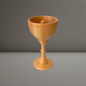 Könnte beinhalten: Ein goldfarbener Kelch mit rundem Fuß und schlankem Stiel. Die Schale hat ein dekoratives Band um den Rand und eine kleine Kugel in der Mitte. Der Kelch besteht aus einem festen Material und hat eine glatte Oberfläche.