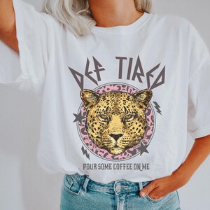 May include: White t-shirt with a graphic of a leopard's face, the words "DEF TIRED" above, and "POUR SOME COFFEE ON ME" below. The leopard graphic is surrounded by a pink circle with stars and lightning bolts.