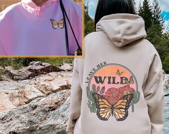 May include: A light beige hoodie with a graphic print of a butterfly and the text "Leave Her Wild" in a circle with a sunset background. The hoodie has a drawstring hood and a kangaroo pocket.