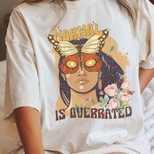 May include: A white t-shirt with a graphic design of a woman with a butterfly over her face. The text "NORMAL IS OVERRATED" is printed below the graphic.