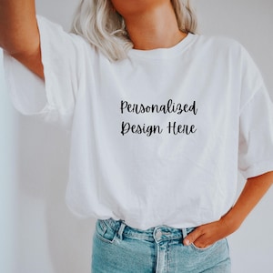 May include: A white t-shirt with the text "Personalized Design Here" printed on the front. The shirt is being worn by a person with blonde hair and blue jeans.