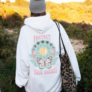 May include: White hooded sweatshirt with a graphic print of a butterfly, sun, moon, and the text "Protect Your Energy!" on the back. The sweatshirt is being worn by a person with a gray beanie and a leopard print tote bag.