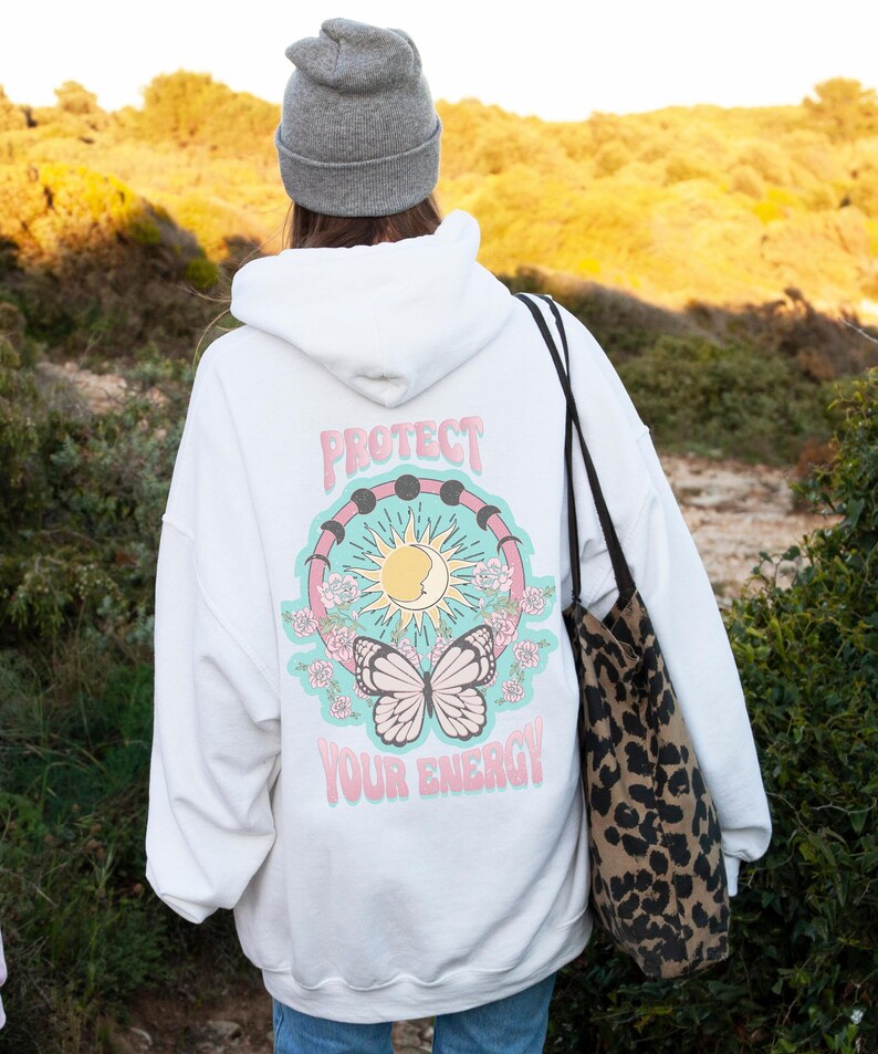 May include: White hooded sweatshirt with a graphic print of a butterfly, sun, moon, and the text "Protect Your Energy!" on the back. The sweatshirt is being worn by a person with a gray beanie and a leopard print tote bag.