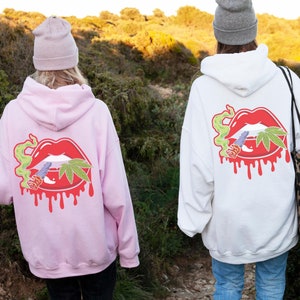May include: Two people wearing pink and white hoodies featuring a graphic of a red, dripping, cartoon mouth with a marijuana leaf and a joint on it. Both hoodies have a hood and a pouch pocket on the front.