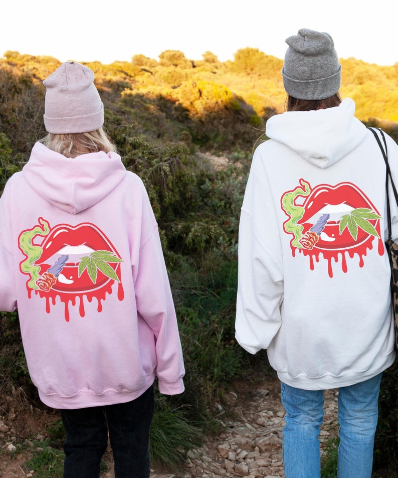 May include: Two people wearing pink and white hoodies featuring a graphic of a red, dripping, cartoon mouth with a marijuana leaf and a joint on it. Both hoodies have a hood and a pouch pocket on the front.