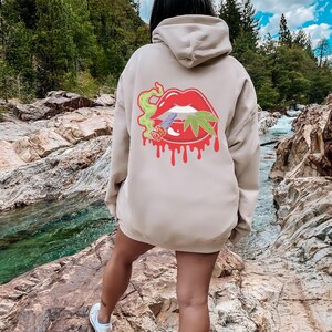 May include: A beige hoodie featuring a graphic of a red dripping mouth with a marijuana leaf and a cigarette inside. The hoodie is being worn by a person standing in a rocky riverbed.