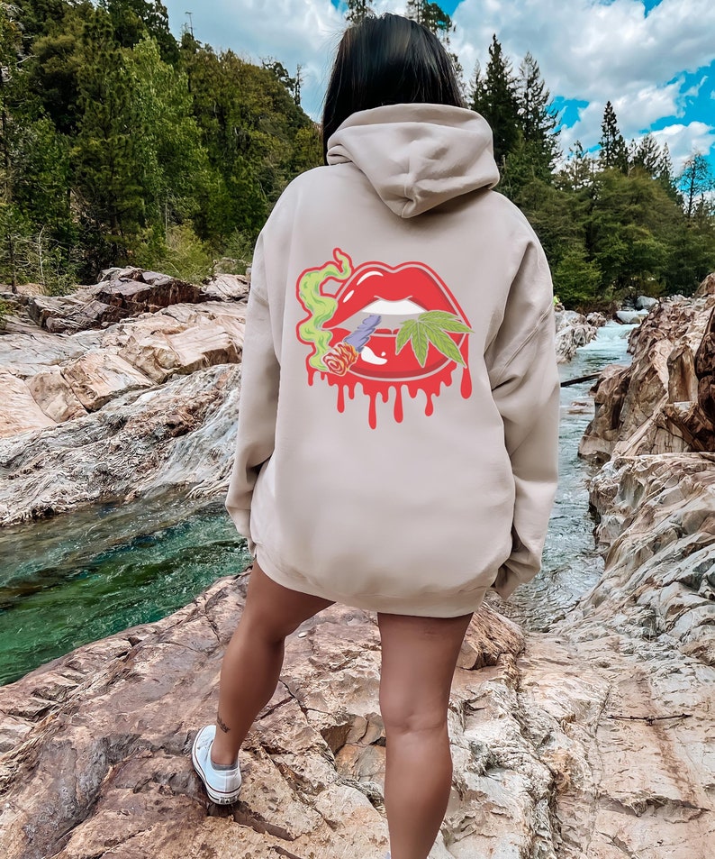 May include: A beige hoodie featuring a graphic of a red dripping mouth with a marijuana leaf and a cigarette inside. The hoodie is being worn by a person standing in a rocky riverbed.