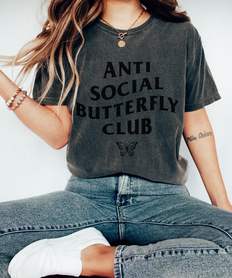 May include: A dark gray t-shirt with the text "Anti Social Butterfly Club" printed in black. A small black butterfly graphic is below the text.