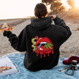 May include: Black hooded sweatshirt featuring a graphic of a red mouth with a marijuana leaf and a joint sticking out. The mouth is dripping red.