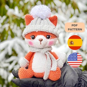 Puede incluir: Amigurumi de zorro de crochet con un gorro gris con un pompón blanco y una bufanda gris. El zorro está sentado en una mano con un fondo nevado. La imagen incluye el texto "PDF PATTERN" en un rectángulo de color melocotón.