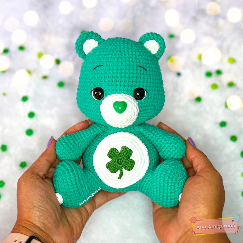 May include: Green crocheted Care Bear with a four-leaf clover on its tummy. The bear is being held by a person.