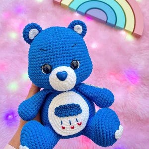 May include: Blue crocheted Care Bear with a white belly and a cloud with raindrops on it. The bear has a blue heart nose and black eyes.
