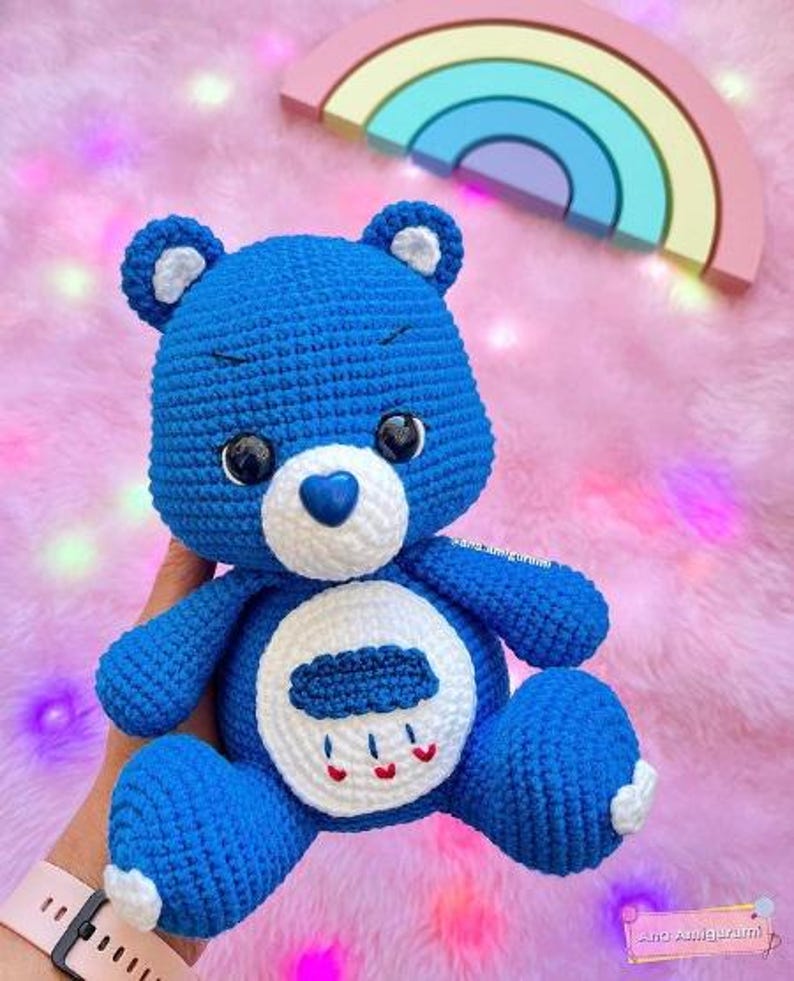 May include: Blue crocheted Care Bear with a white belly and a cloud with raindrops on it. The bear has a blue heart nose and black eyes.