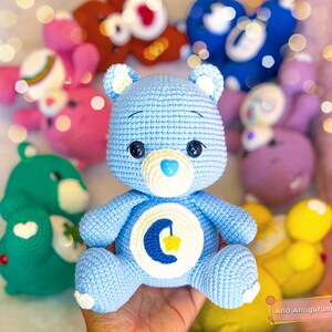 May include: A light blue crocheted Care Bear with a crescent moon and star on its tummy. The bear has a white heart-shaped nose and black eyes. The bear is surrounded by other crocheted Care Bears in various colours.