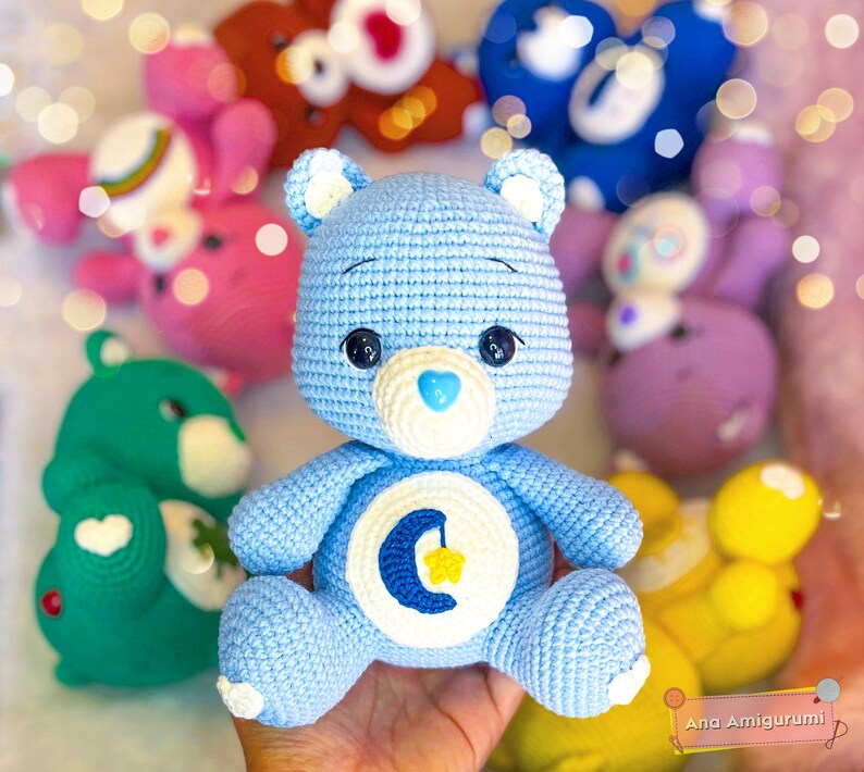 May include: A light blue crocheted Care Bear with a crescent moon and star on its tummy. The bear has a white heart-shaped nose and black eyes. The bear is surrounded by other crocheted Care Bears in various colours.