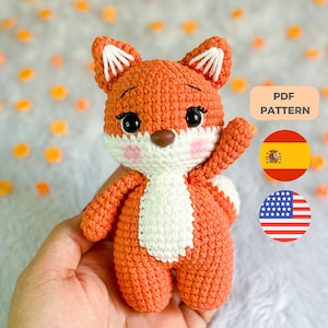 Puede incluir: Un zorro naranja tejido a ganchillo con detalles blancos, ojos negros y mejillas rosadas. El zorro levanta una pata. La imagen incluye el texto "PDF PATTERN" y las banderas de España y Estados Unidos.