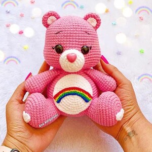 May include: A pink crocheted Care Bear with a rainbow on its chest. The bear has a white face with brown eyes and a pink nose.