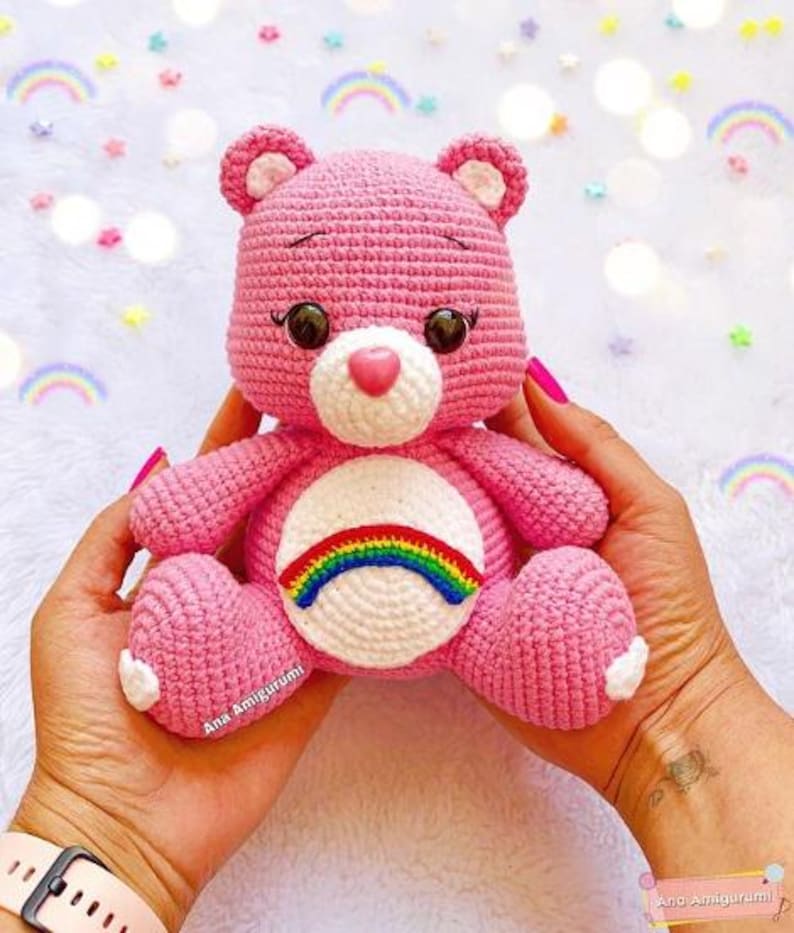 May include: A pink crocheted Care Bear with a rainbow on its chest. The bear has a white face with brown eyes and a pink nose.