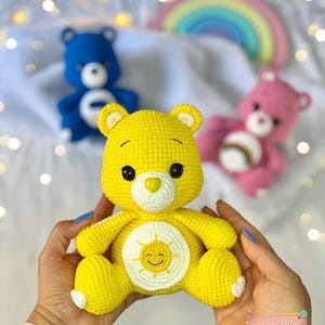 May include: A yellow crocheted Care Bear with a smiling sun on its tummy. The bear is being held by a person's hands.