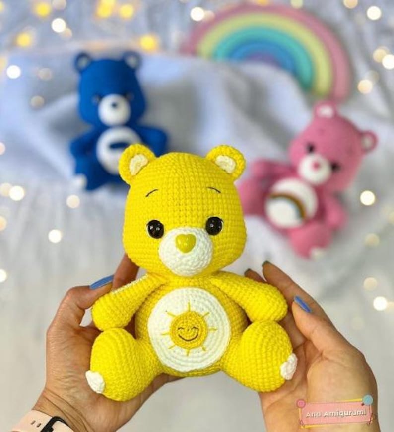 May include: A yellow crocheted Care Bear with a smiling sun on its tummy. The bear is being held by a person's hands.