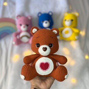 May include: A brown crocheted teddy bear with a white heart on its chest. The heart has a smaller red heart in the centre. The bear is sitting on a hand.