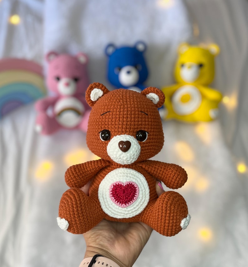 May include: A brown crocheted teddy bear with a white heart on its chest. The heart has a smaller red heart in the centre. The bear is sitting on a hand.