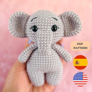 Pode incluir: Um pequeno elefante de crochê cinza com orelhas grandes e olhos pretos. O elefante feito à mão tem uma aparência amigável. A imagem inclui um rótulo "PDF PATTERN" e bandeiras da Espanha e dos Estados Unidos.