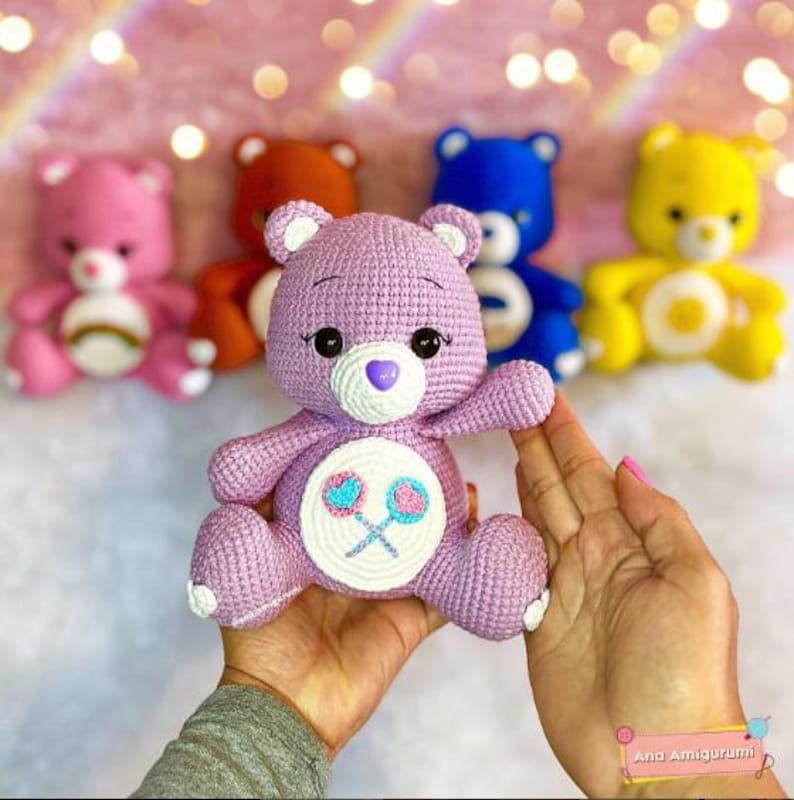 May include: A purple crocheted Care Bear with a white belly and a design of two pink and blue lollipops. The bear has a purple nose and a white heart on its chest.