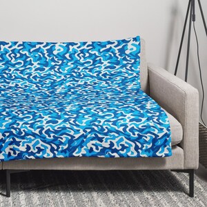 May include: A blue and white patterned fleece throw blanket draped over a gray couch. The blanket has a modern abstract design.