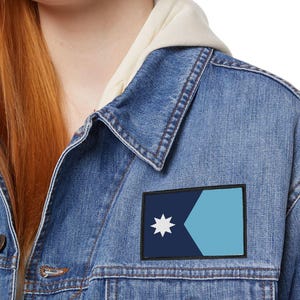 May include: A denim jacket with a patch featuring a navy blue and light blue geometric design and a white star. The jacket has a classic button-up design and is worn over a cream-colored hoodie.