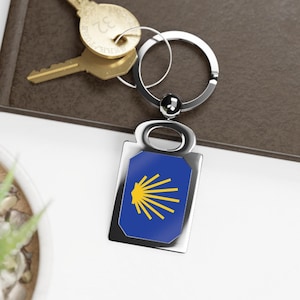 Puede incluir: Un llavero de plata con un dije de concha de vieira azul y amarillo. El dije es un símbolo del Camino de Santiago.
