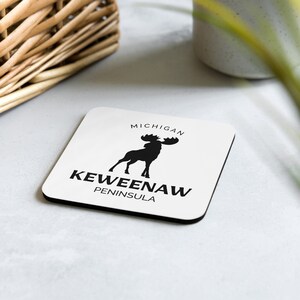 May include: White square coaster with black text and a black silhouette of a moose. The text reads "MICHIGAN KEWEENAW PENINSULA".