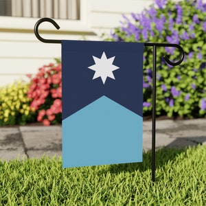 Könnte beinhalten: Eine blaue und hellblaue Gartenflagge mit einem weißen Stern in der Mitte. Die Flagge ist an einer schwarzen Metallstange befestigt.
